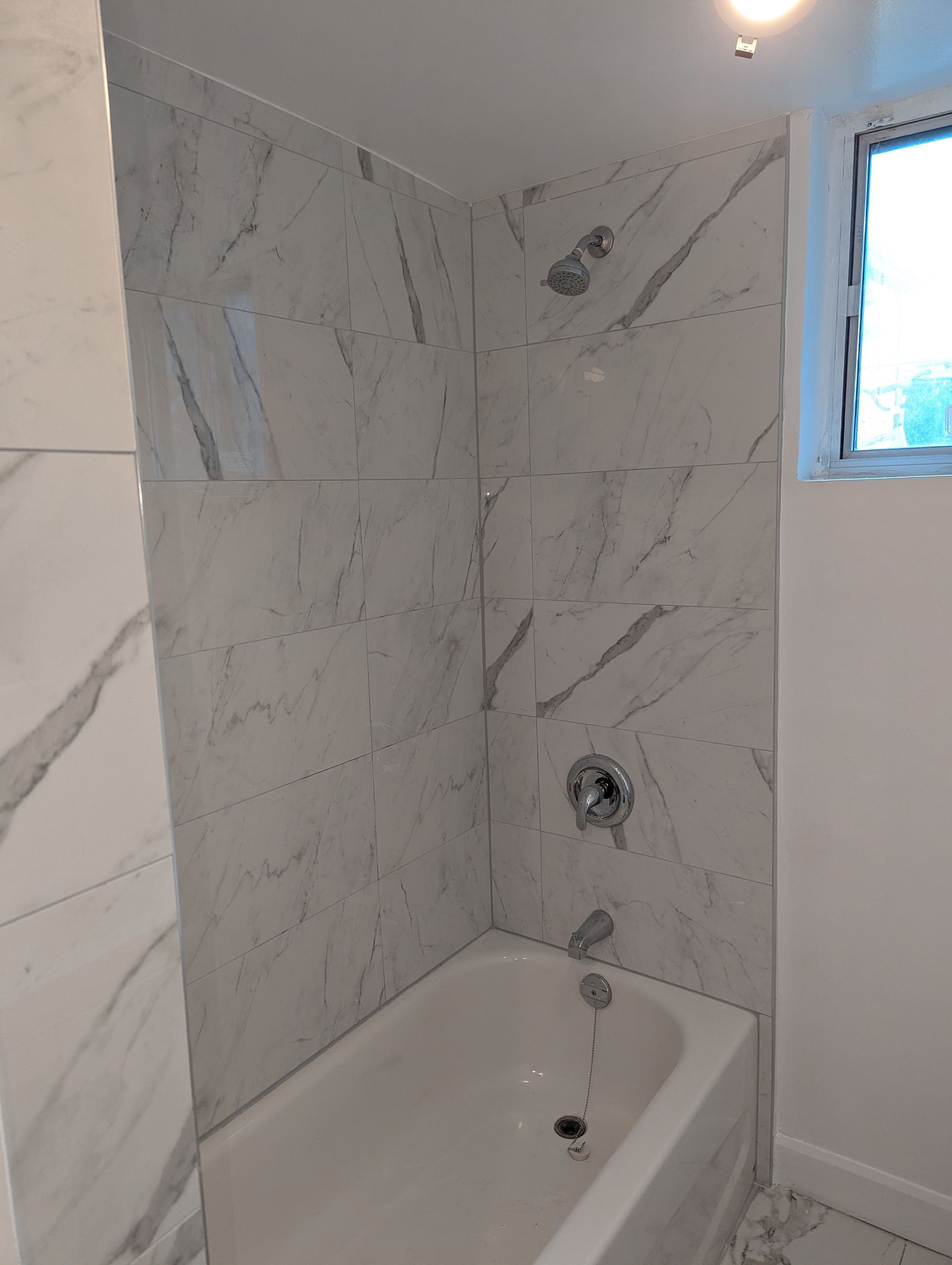 Small bathroom with a white bathtub, marble-pattern tile walls, and a window above.