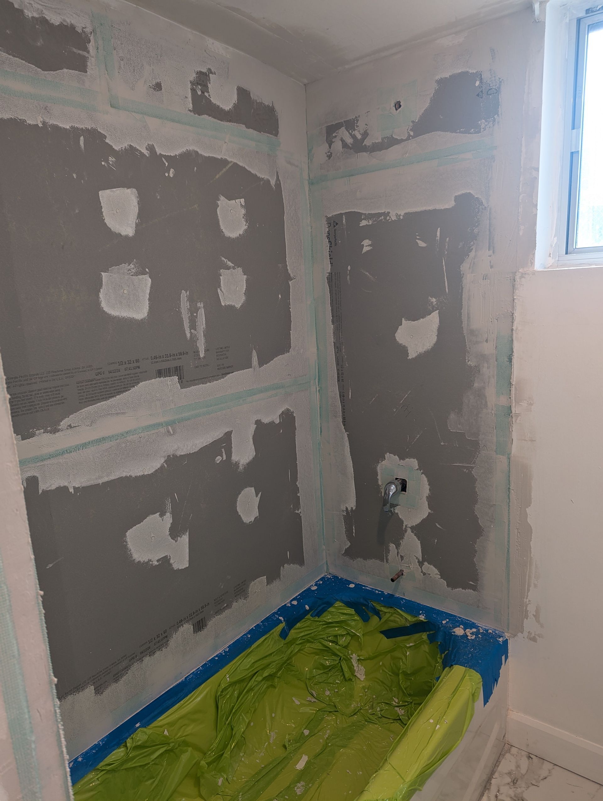 Small bathroom under renovation with gray patched drywall, blue tape, and a covered floor corner