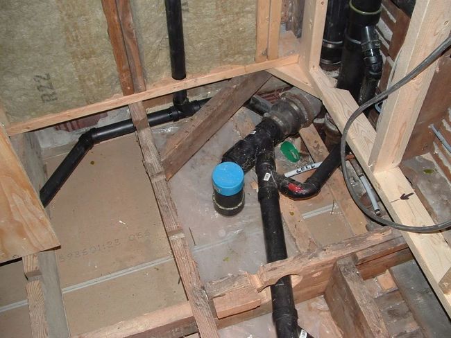 Exposed plumbing under a floor, with a blue-capped pipe and wiring in a construction opening