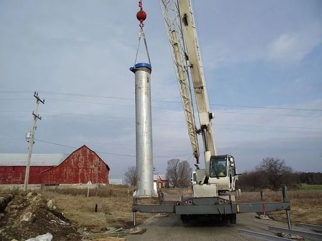 Crane lifting a tall silver metal pole beside a red barn in a rural field.