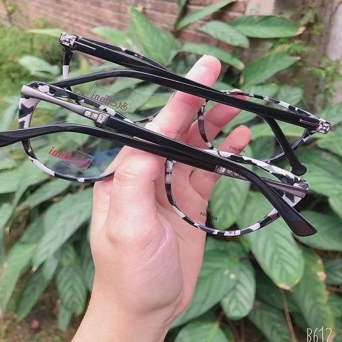 Unas gafas rectangulares negras con cristales transparentes, sostenidas en una mano sobre un fondo de hojas verdes.