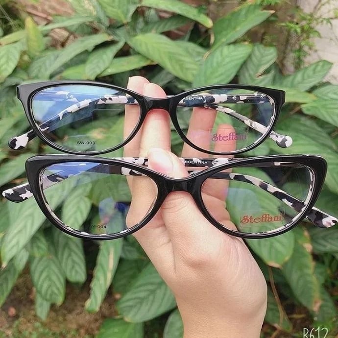 Dos monturas de gafas negras sostenidas en una mano contra hojas verdes