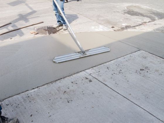 A worker smooths wet concrete on a pavement using a long metal tool.