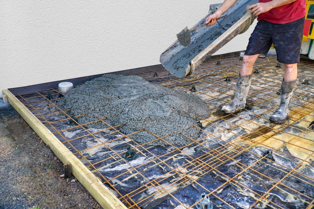 A construction worker in boots and shorts pours wet concrete onto a rebar grid for a foundation.