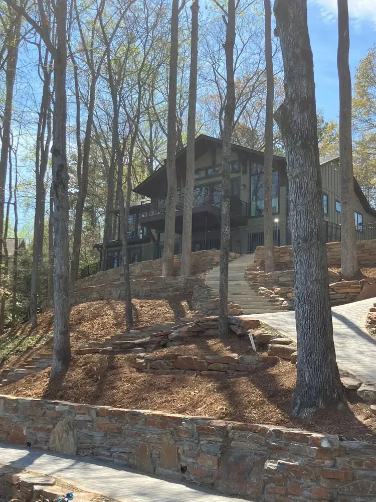 Wooded hillside with a stone house, tall trees, and a stone retaining wall under a clear sky