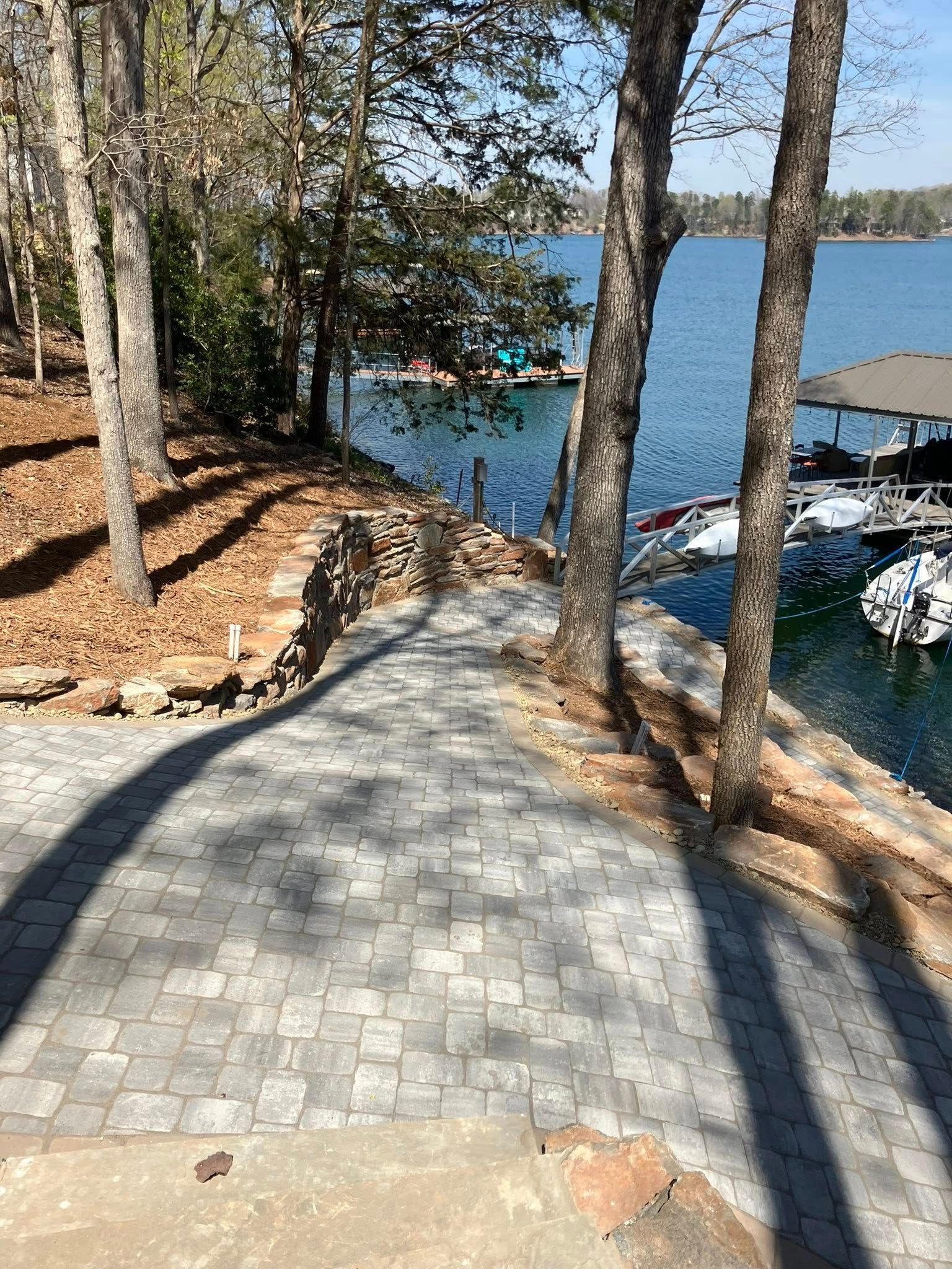 Stone path through trees leading down to a blue lake with docked boats and a small shoreline.