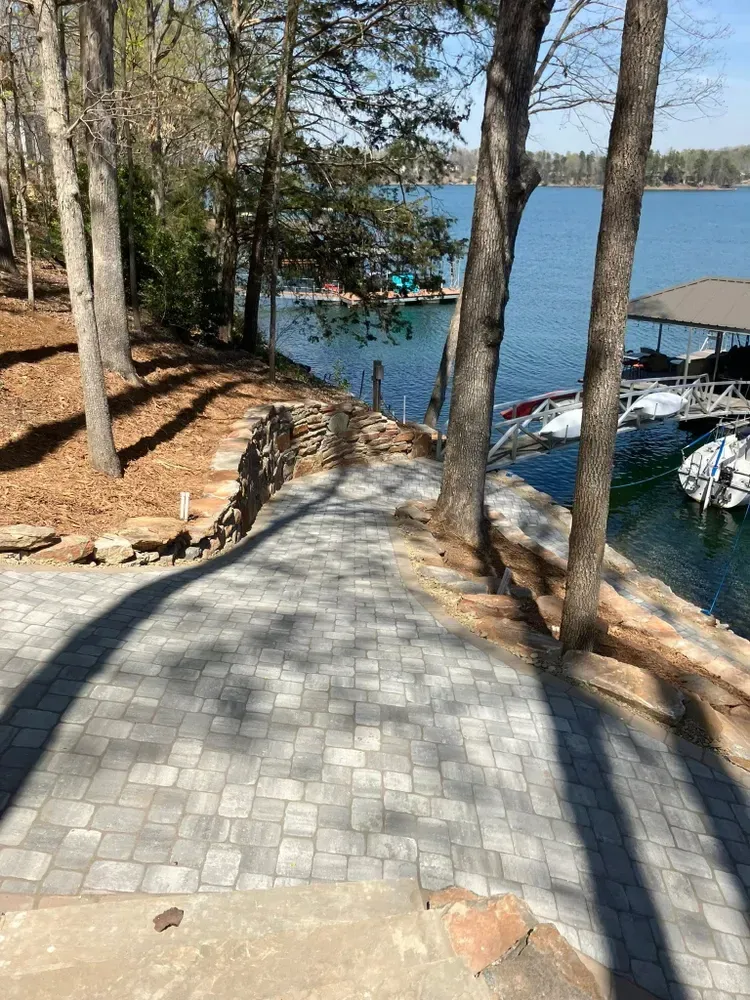 Stone path through trees leading to a lakeside dock with boats and blue water.