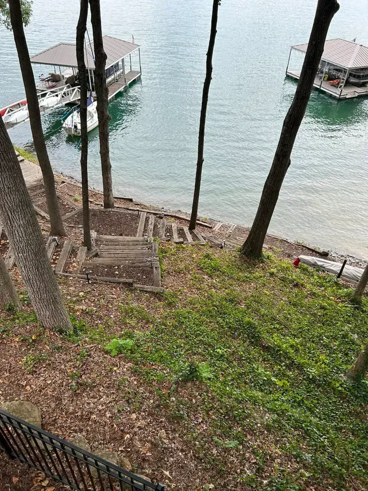 Steep lakeside path with stone steps and railings, overlooking calm water and docked boats between tall trees.