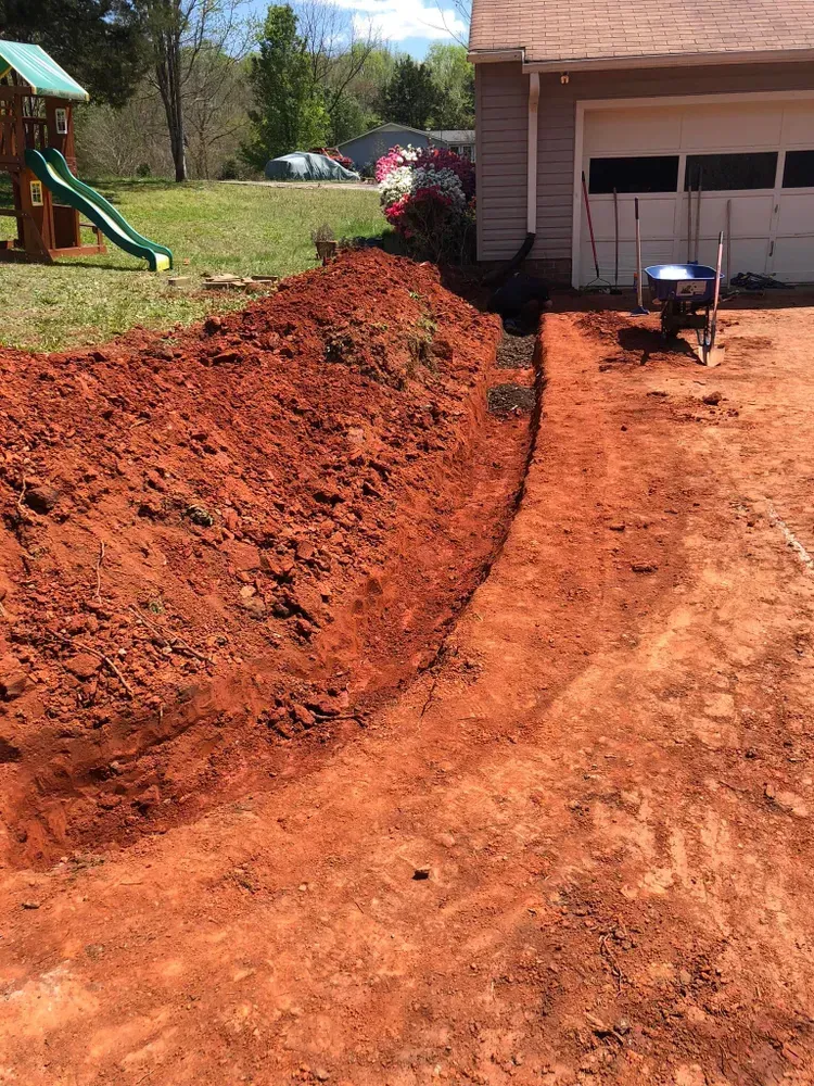 Fresh red dirt piled along a driveway beside a garage, with a wheelbarrow on the right and a playground on the left