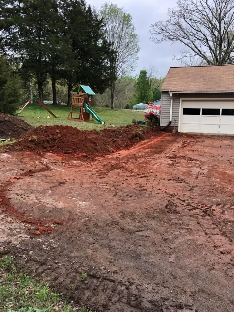 Red dirt driveway excavation beside a garage and playground in a wooded yard