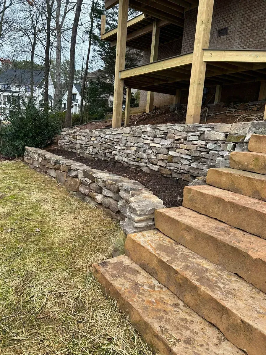 Stone retaining wall and wooden stairs beside a house on a grassy slope in a wooded yard