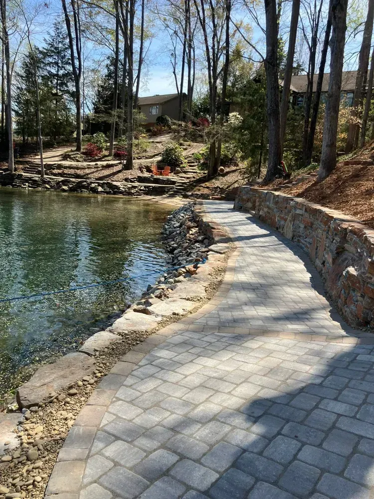 Curved stone walkway beside a pond, with trees, shoreline rocks, and houses in the background