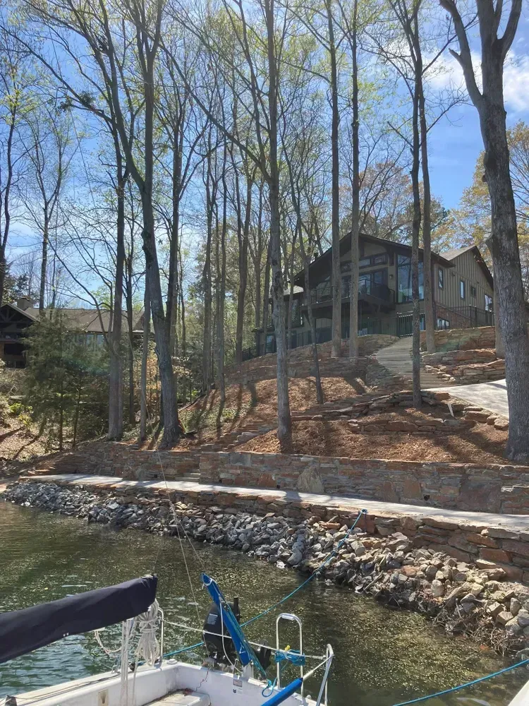 Wooded lakeside with a house on a hill, stone retaining wall, and docked boats in the foreground