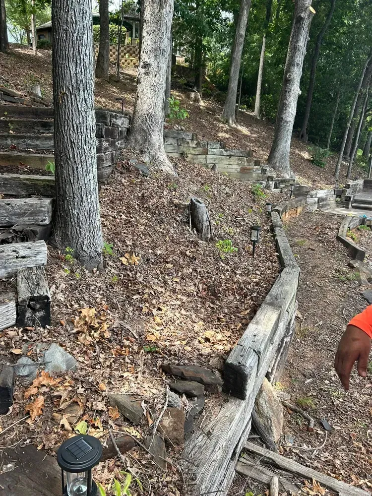 Rocky wooded hillside with trees, a narrow dirt path, and a weathered wooden barrier beside a fence