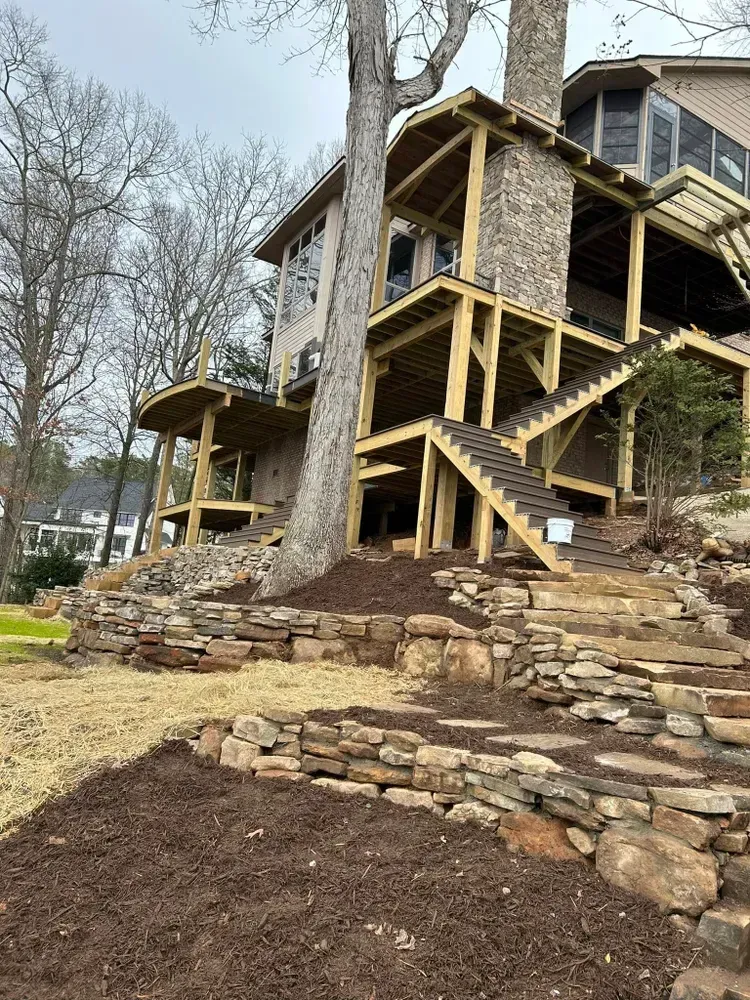 Stone house with wooden stairs and decks on a wooded hillside, with a tree growing beside it