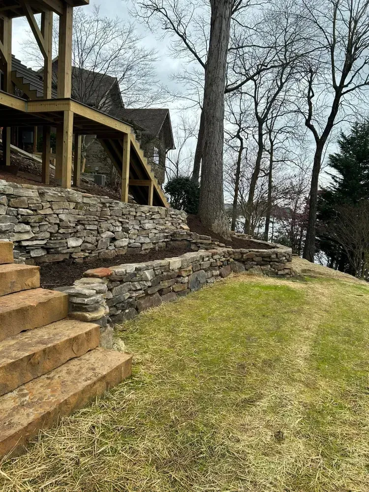 Stone steps and retaining wall beside a grassy slope and leafless trees next to a wooden deck