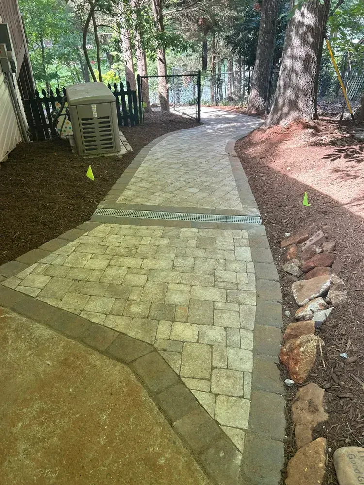 Paved walkway under trees beside a house, with a fence and leaf-covered ground.