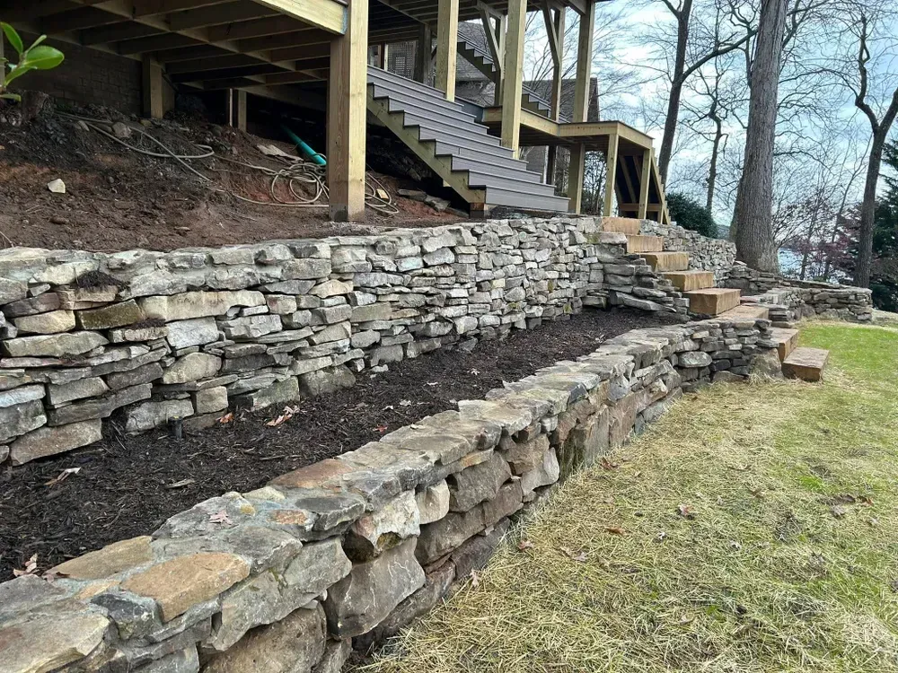 Stone retaining wall with stepped path and wooden staircase under a deck in a wooded yard