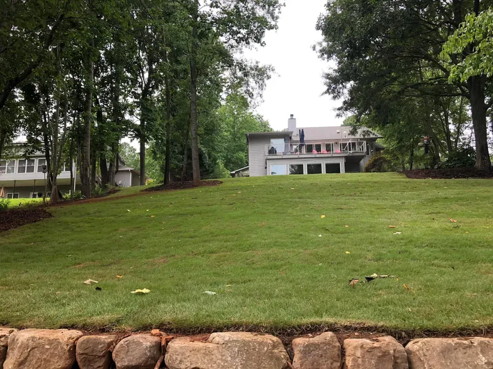 Grassy hill with a stone edge, trees, and a white house with flags in the background.