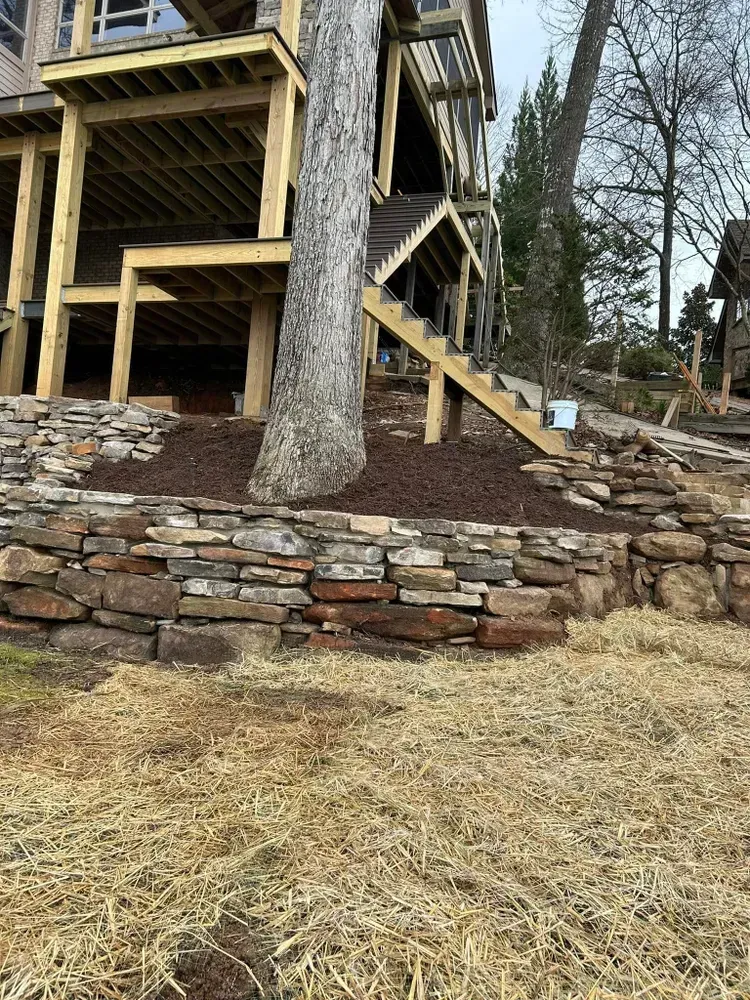 Stone retaining wall and wooden stairway beside a raised house under construction in a wooded yard.