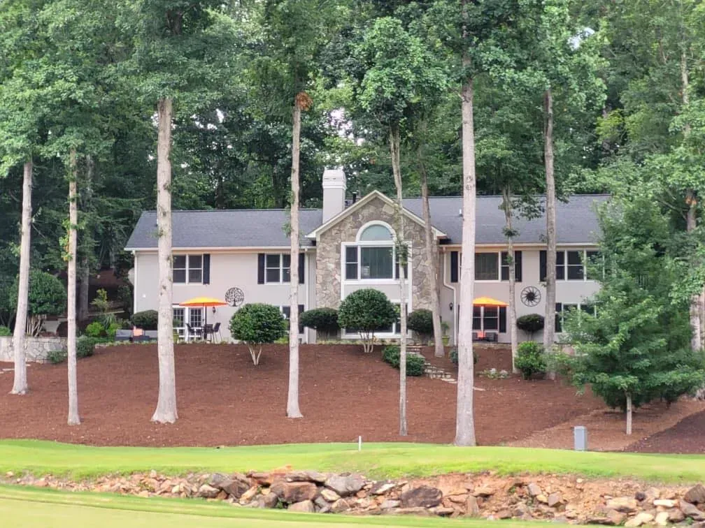 Large white house with gray roof and stone entry, surrounded by tall trees and pine-covered landscaping.