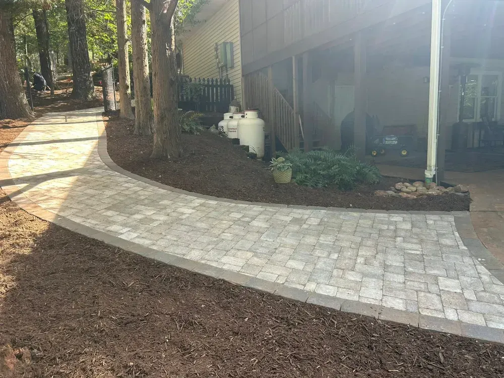 Curved light stone walkway beside a house, bordered by mulch and trees in a wooded yard.