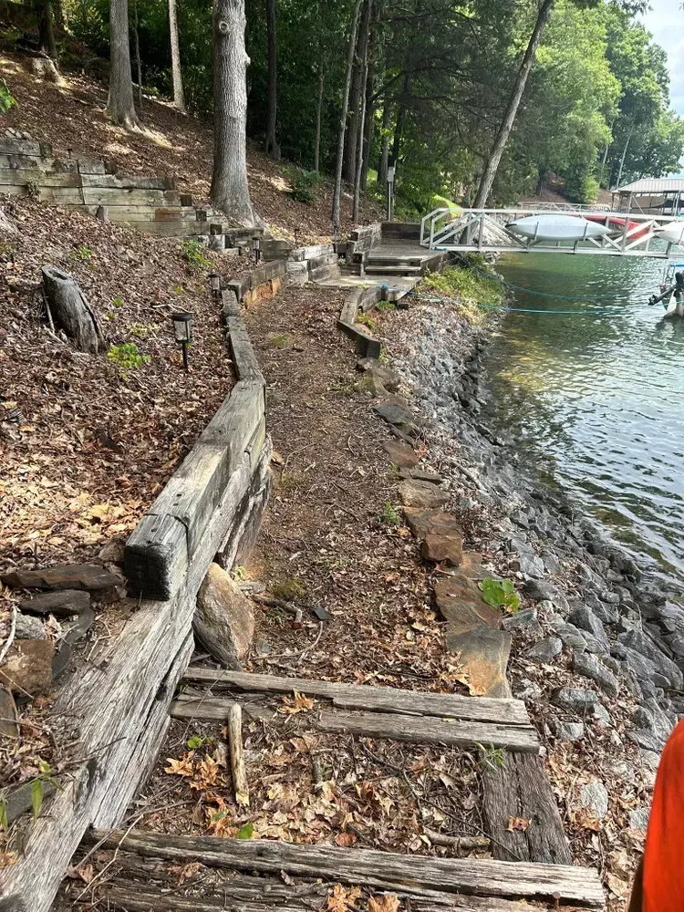 Eroded lakeshore with wooden retaining timbers, trees on the left, and a dock with boats in the distance