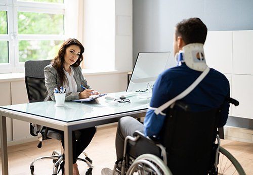 A man in a wheelchair is sitting at a desk talking to a woman.