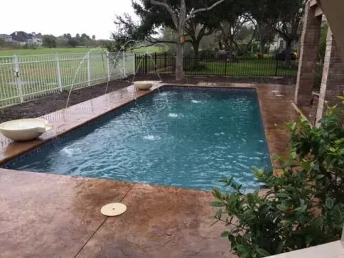 A large swimming pool is surrounded by a fence and trees in a backyard.