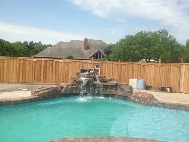 A wooden fence surrounds a swimming pool with a waterfall in the middle.