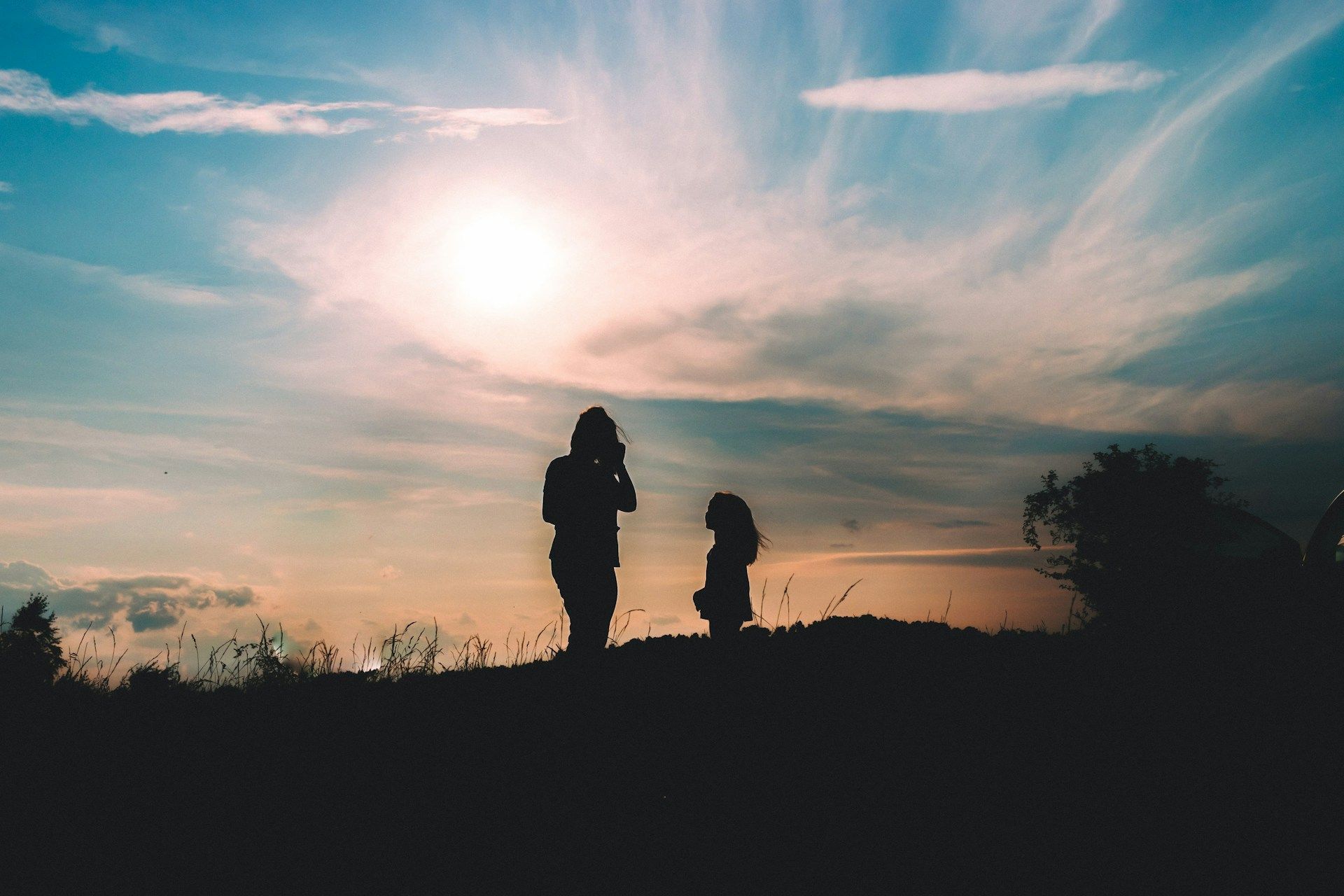 Silhouettes of a woman and a child version of herself on a hill against a vibrant, hazy sunset sky.