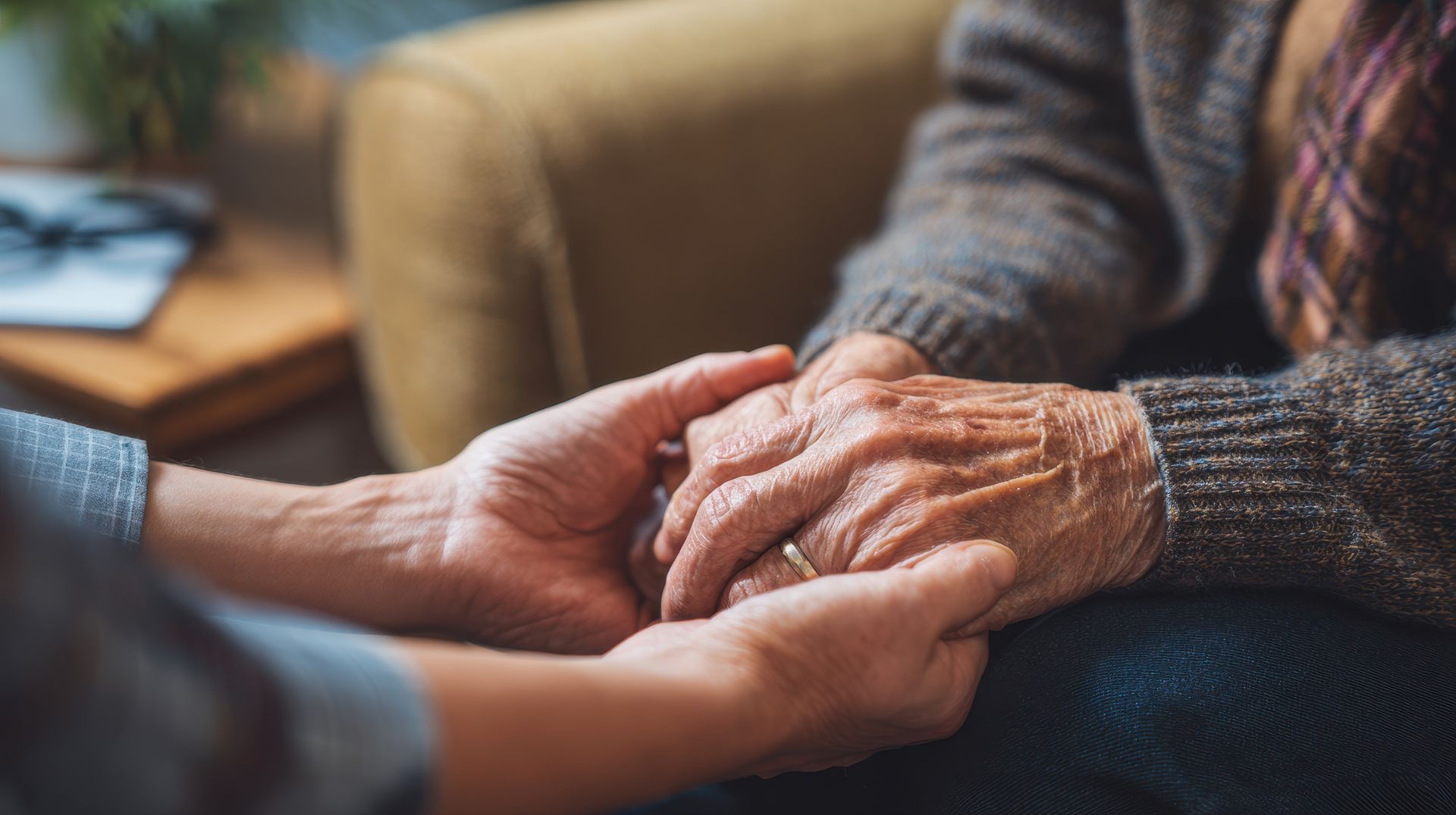 Person holding the weathered hands of another, indoors. Comforting gesture, sweater sleeves visible.
