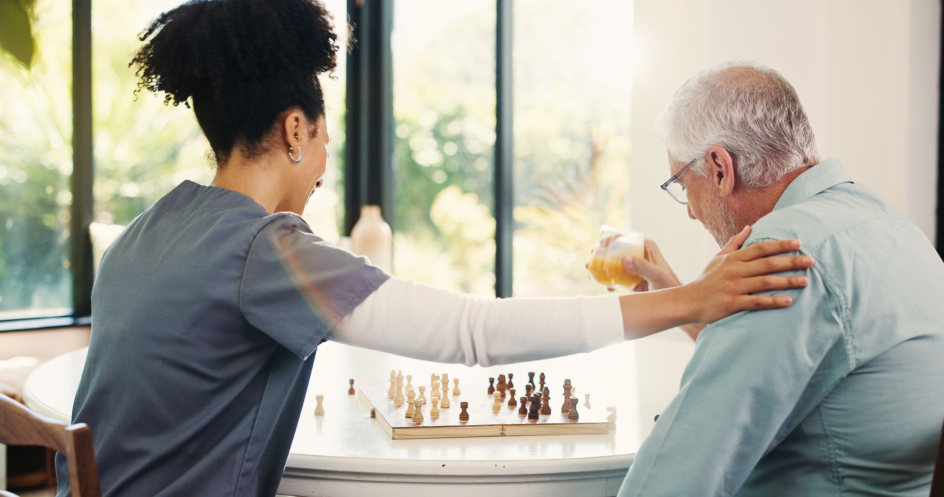 Caregiver assisting an older person, offering a drink while touching their shoulder. Chess board visible.