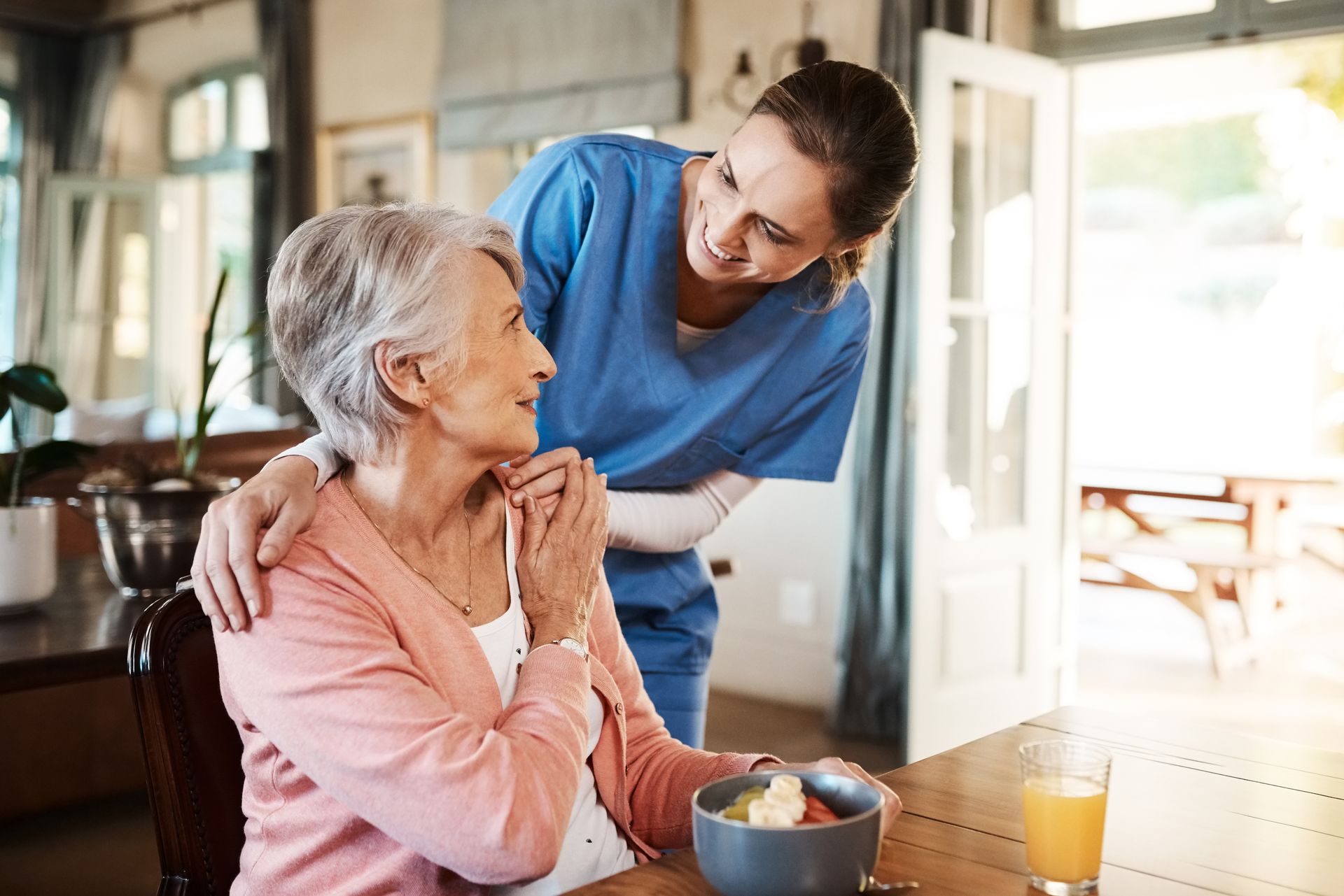 Caregiver smiles at senior woman seated at a table, both indoors.