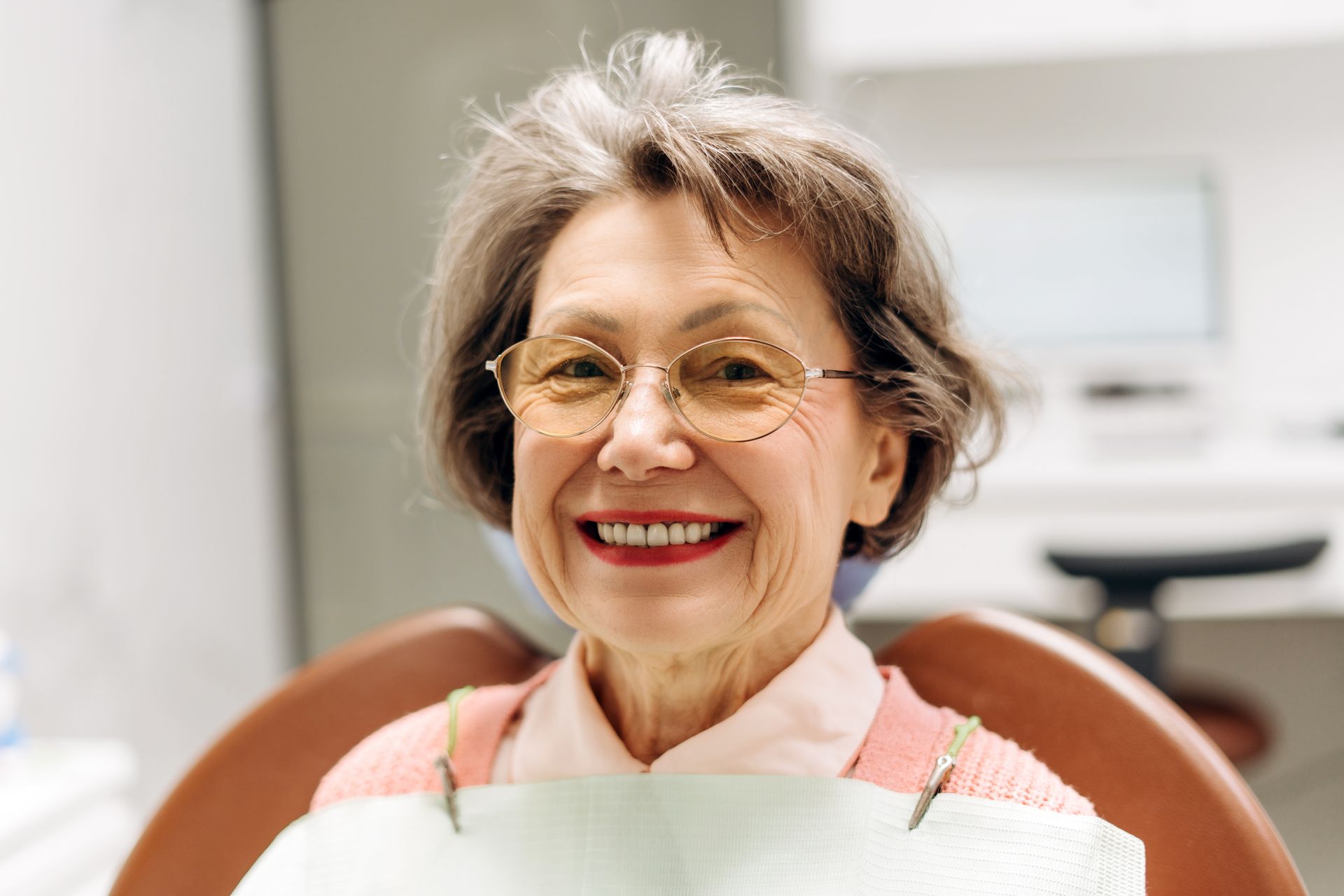 Smiling woman in glasses sitting at a dental chair in a bright clinic