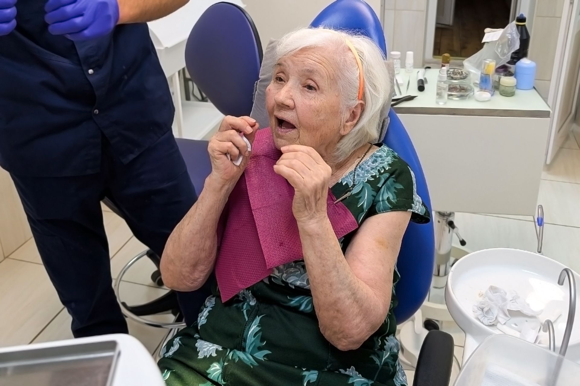 Patient in a dental chair with gloved staff nearby, adjusting a pink bib in a clinic.