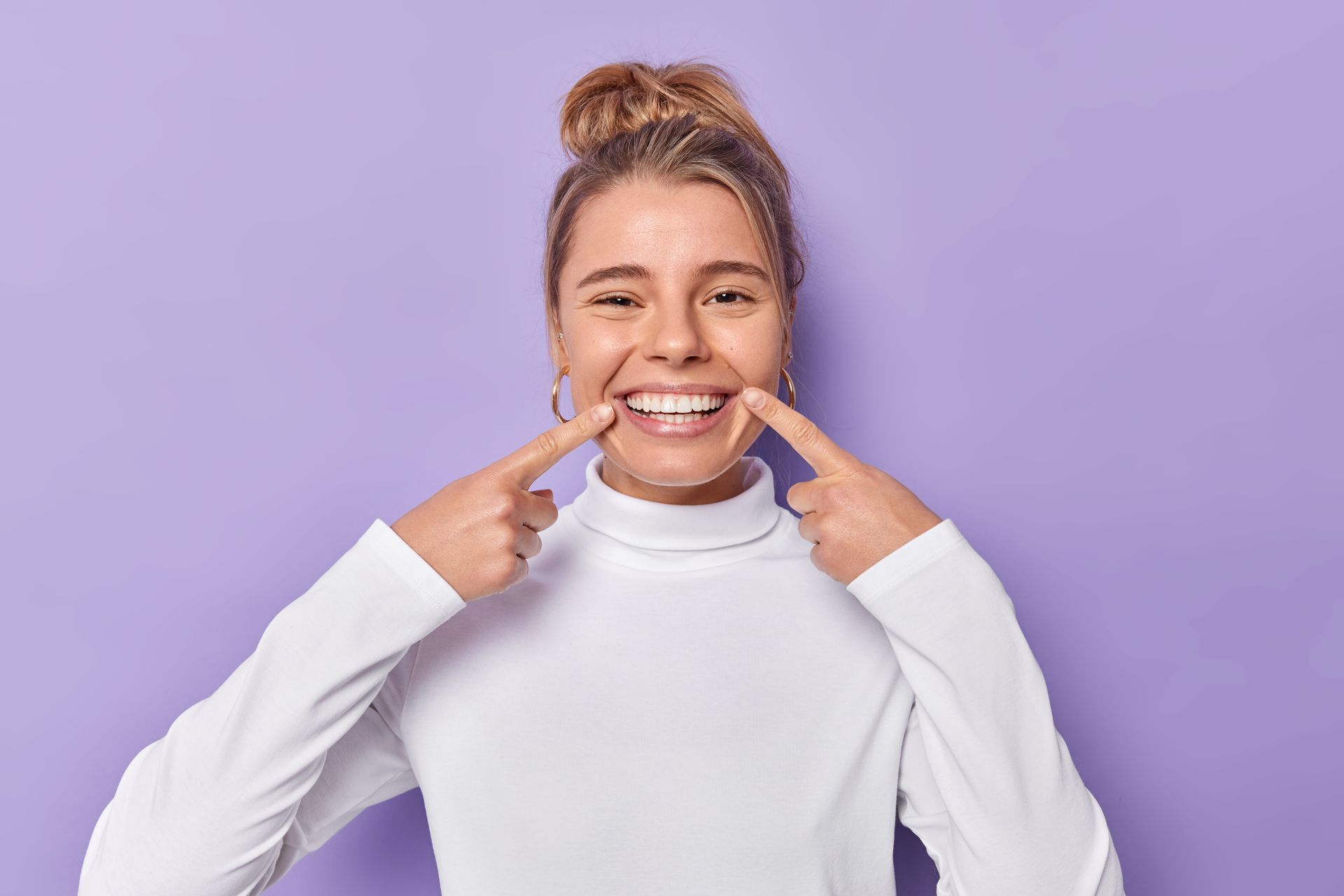 Smiling person in a white top pointing to their teeth against a purple background