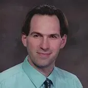 Studio headshot of a man in a light teal shirt and patterned tie, smiling against a dark background