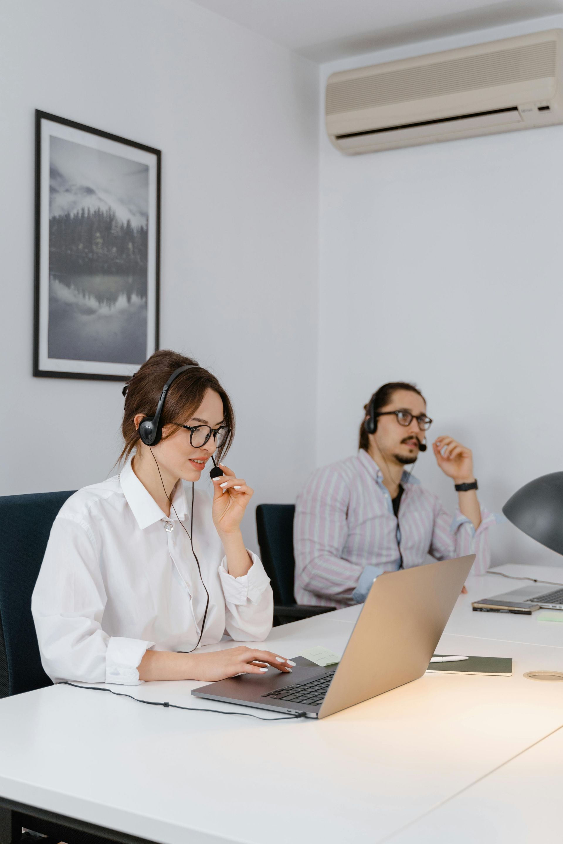 Two office workers wearing headsets work at laptops in a bright meeting room.