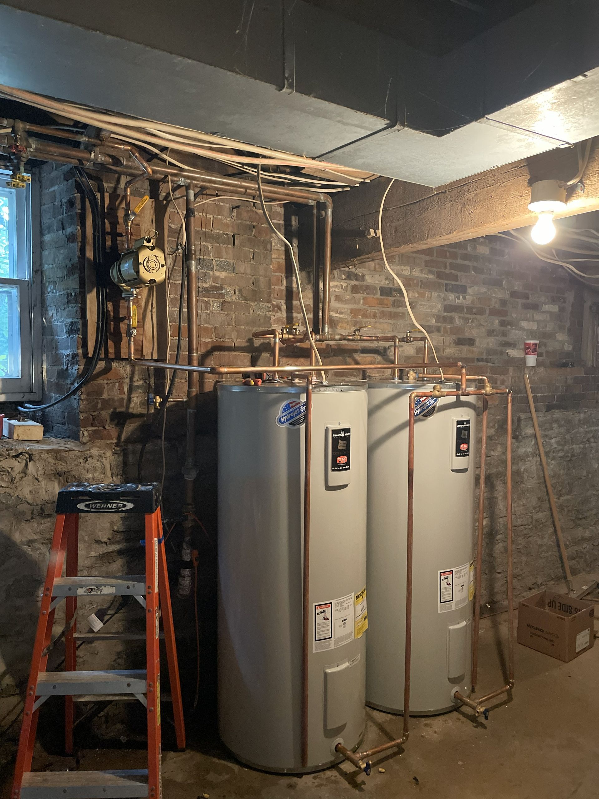 Basement utility area with two water heaters, exposed pipes, and a red ladder under unfinished ceiling.