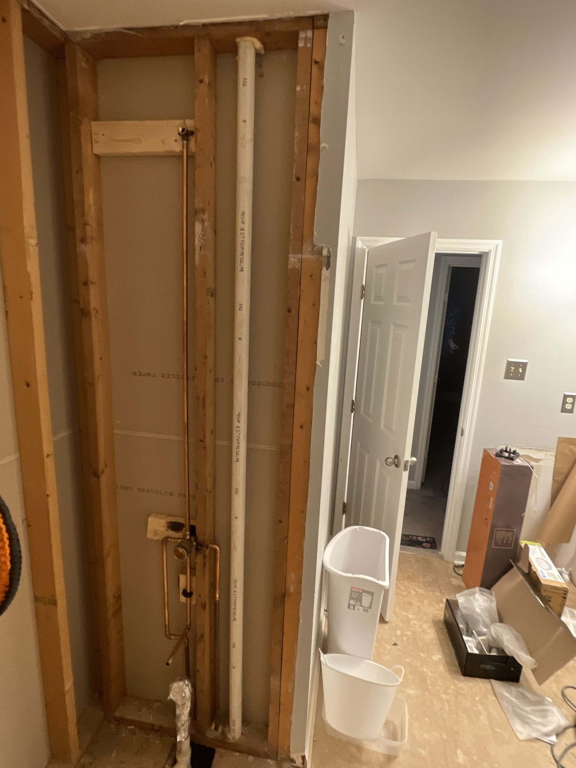 Exposed wall framing and plumbing in a room under renovation, with a white chair and open door.