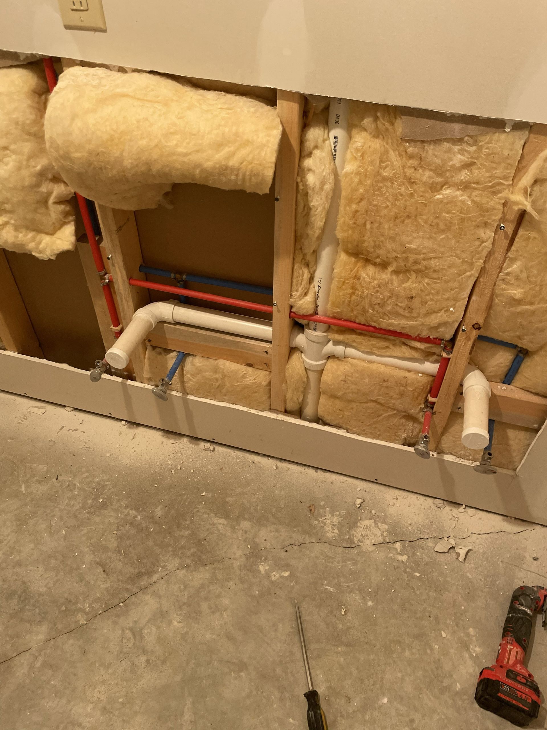 Exposed plumbing and insulation inside an unfinished wall, with concrete floor and a red power tool nearby