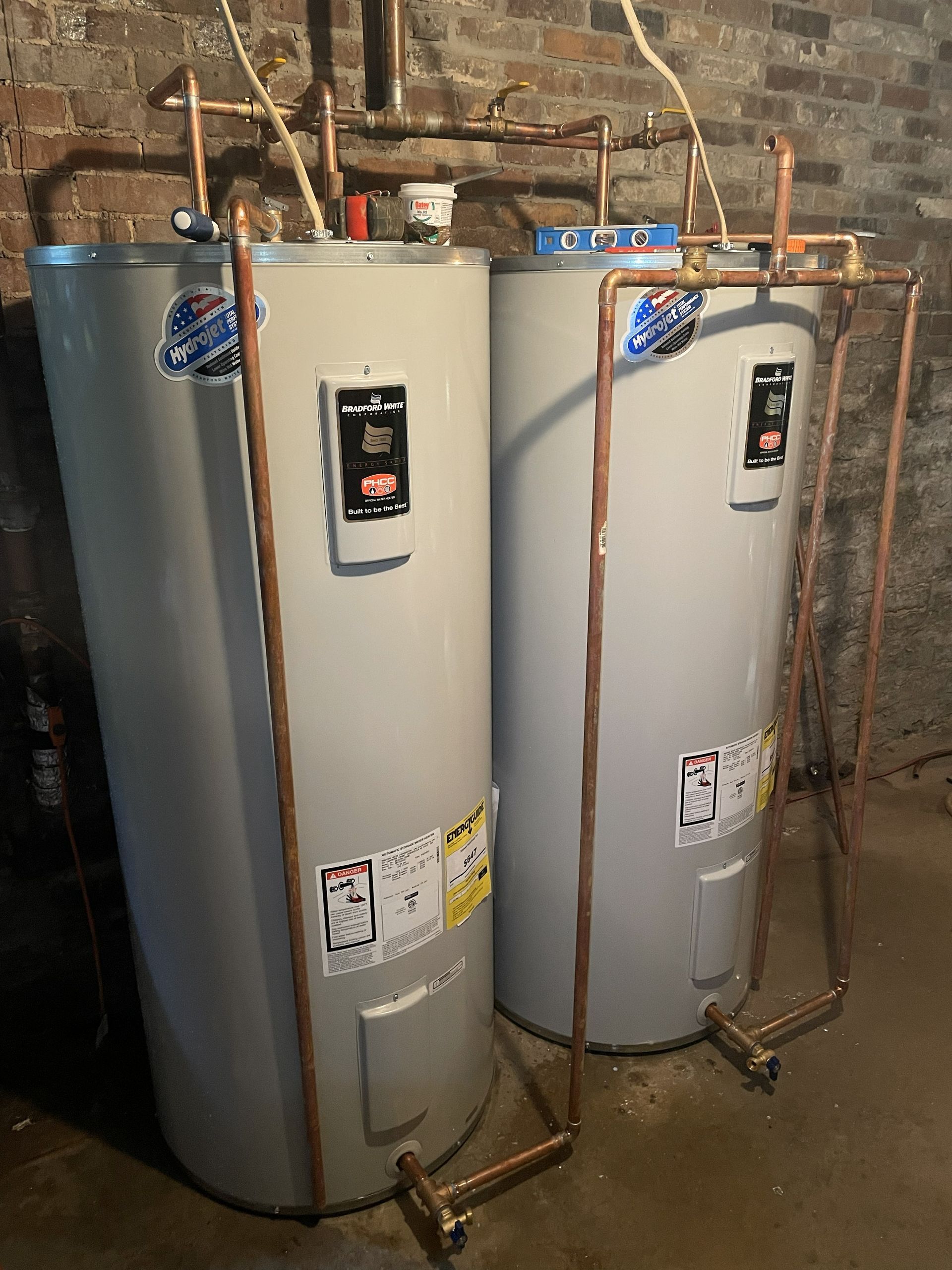 Two tall white water heaters beside a brick wall in a basement