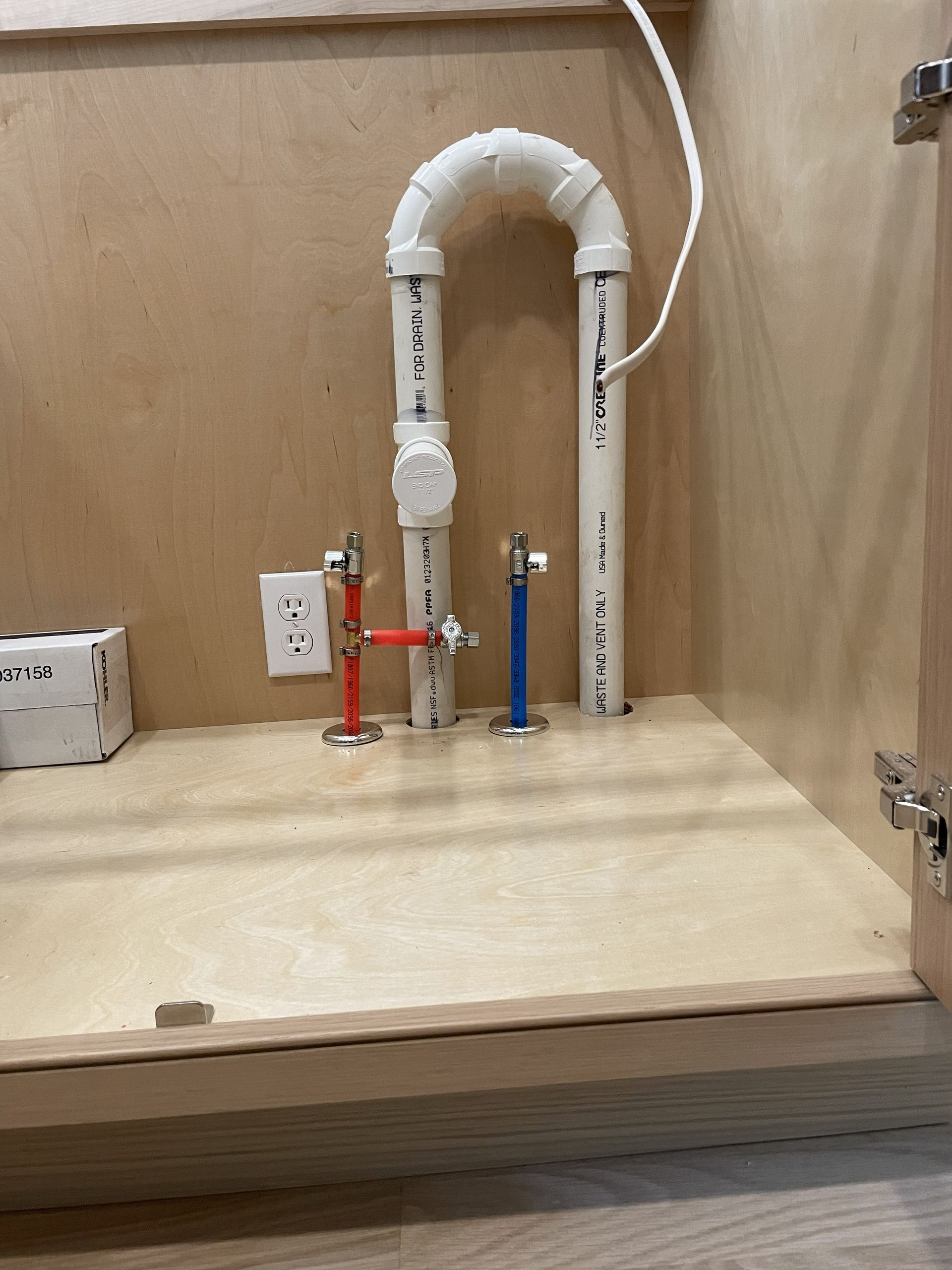 Under-sink plumbing with PVC pipes, shutoff valves, and a cabinet electrical outlet on a wood shelf
