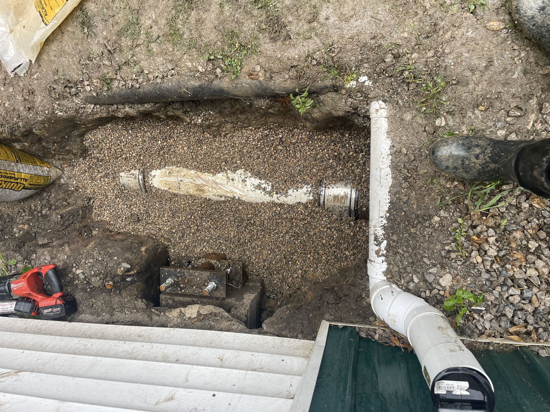 Backyard trench with white drain pipe and elbow beside a house wall and steps, tools nearby
