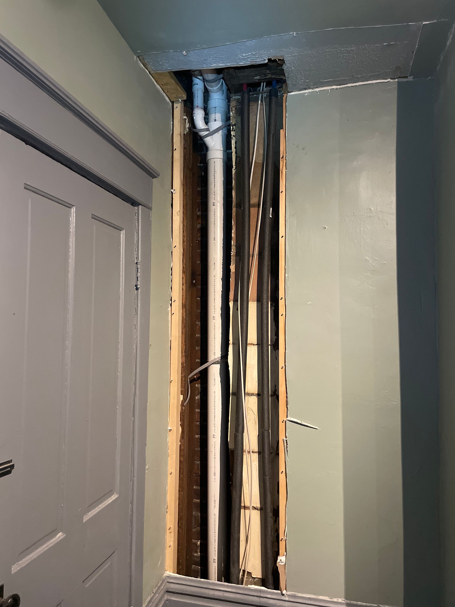 Exposed wall framing and plumbing in a hallway beside a white door, with drywall removed around a vertical opening