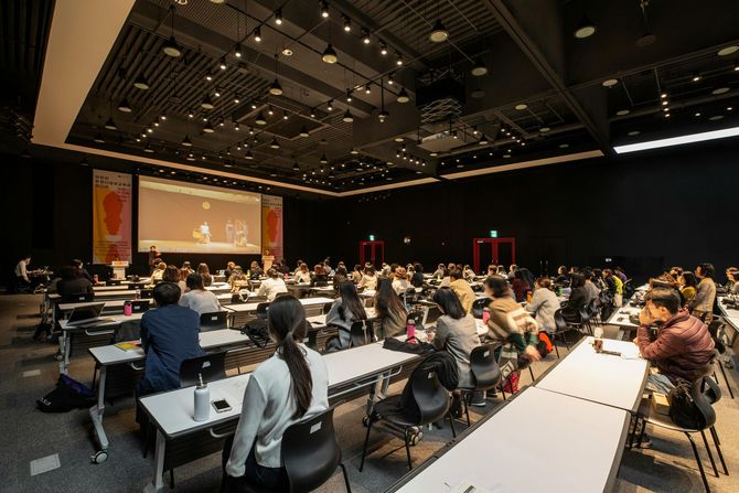 Large conference hall with seated audience facing a stage and projection screen under dim ceiling lights