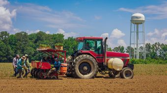 Farmers work beside a red tractor in a field, with a water tower under a blue sky.