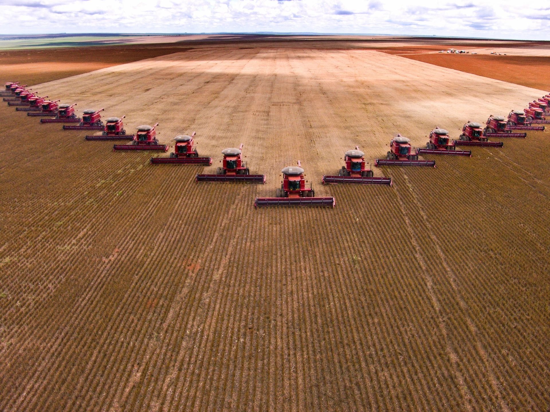 Multiple red harvesters forming a V across a large brown field under a hazy sky