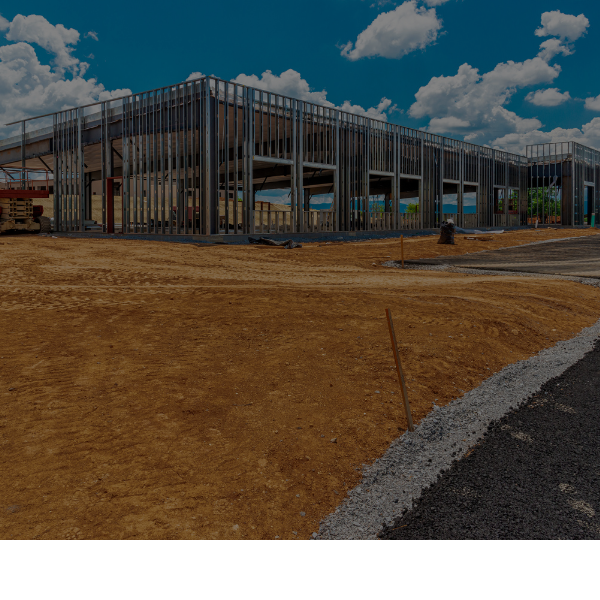 Steel-framed building under construction beside a dirt road and brown grass under a cloudy blue sky